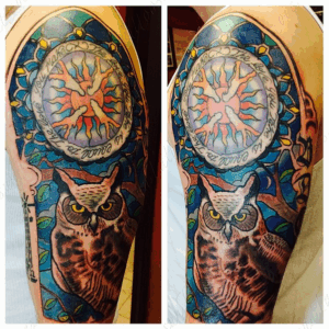 Arm tattoo in color ink of a stained glass design with an owl perched in a tree and a sun with script by Jaesun Duggan