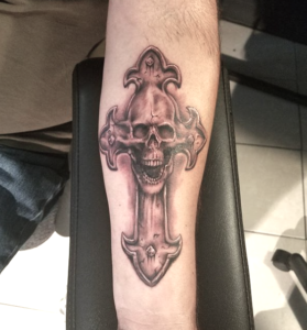 Arm showing tattoo with a skull coming out of a cross in black and gray ink by Kit Marlow
